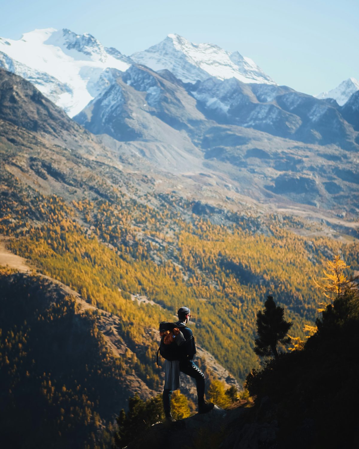 A hiker pausing on a ridge to look out across an autumn valley toward snow-capped peaks